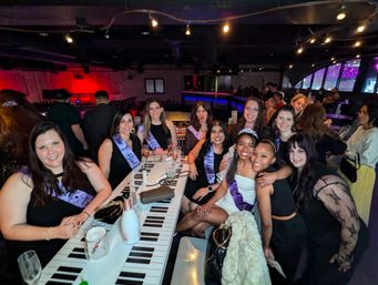 Bachelorette party at a lively indoor lounge — group of friends wearing purple sashes and a tiara, champagne on a piano-key bar table under colorful neon lights.