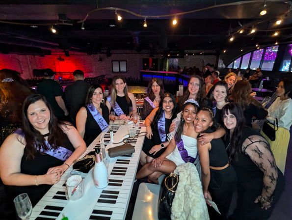 Bachelorette party at a lively indoor lounge — group of friends wearing purple sashes and a tiara, champagne on a piano-key bar table under colorful neon lights.