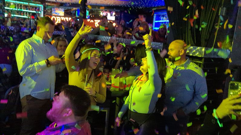 Lively bar party with people dancing and cheering under colorful lights and falling confetti, many holding drinks and raising their arms