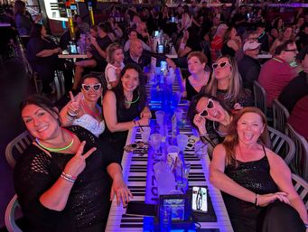 Group of friends laughing and posing at a long piano-key bar table, wearing neon glow necklaces and heart-shaped sunglasses, with drinks and pitchers in a crowded nightlife setting