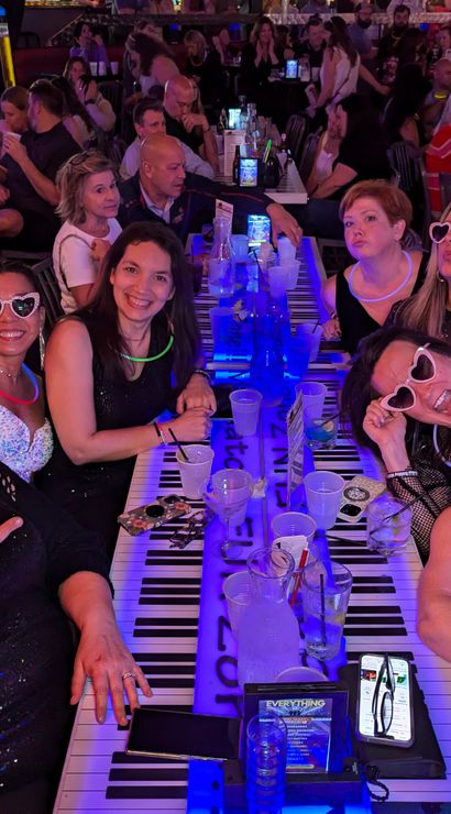 Group of friends laughing and posing at a long piano-key bar table, wearing neon glow necklaces and heart-shaped sunglasses, with drinks and pitchers in a crowded nightlife setting