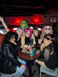 Group of people in colorful wigs and sunglasses posing around a high-top table with drinks and a snack basket in a brick-walled bar under red ambient lighting — lively downtown nightlife scene.