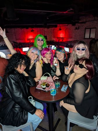 Group of people in colorful wigs and sunglasses posing around a high-top table with drinks and a snack basket in a brick-walled bar under red ambient lighting — lively downtown nightlife scene.