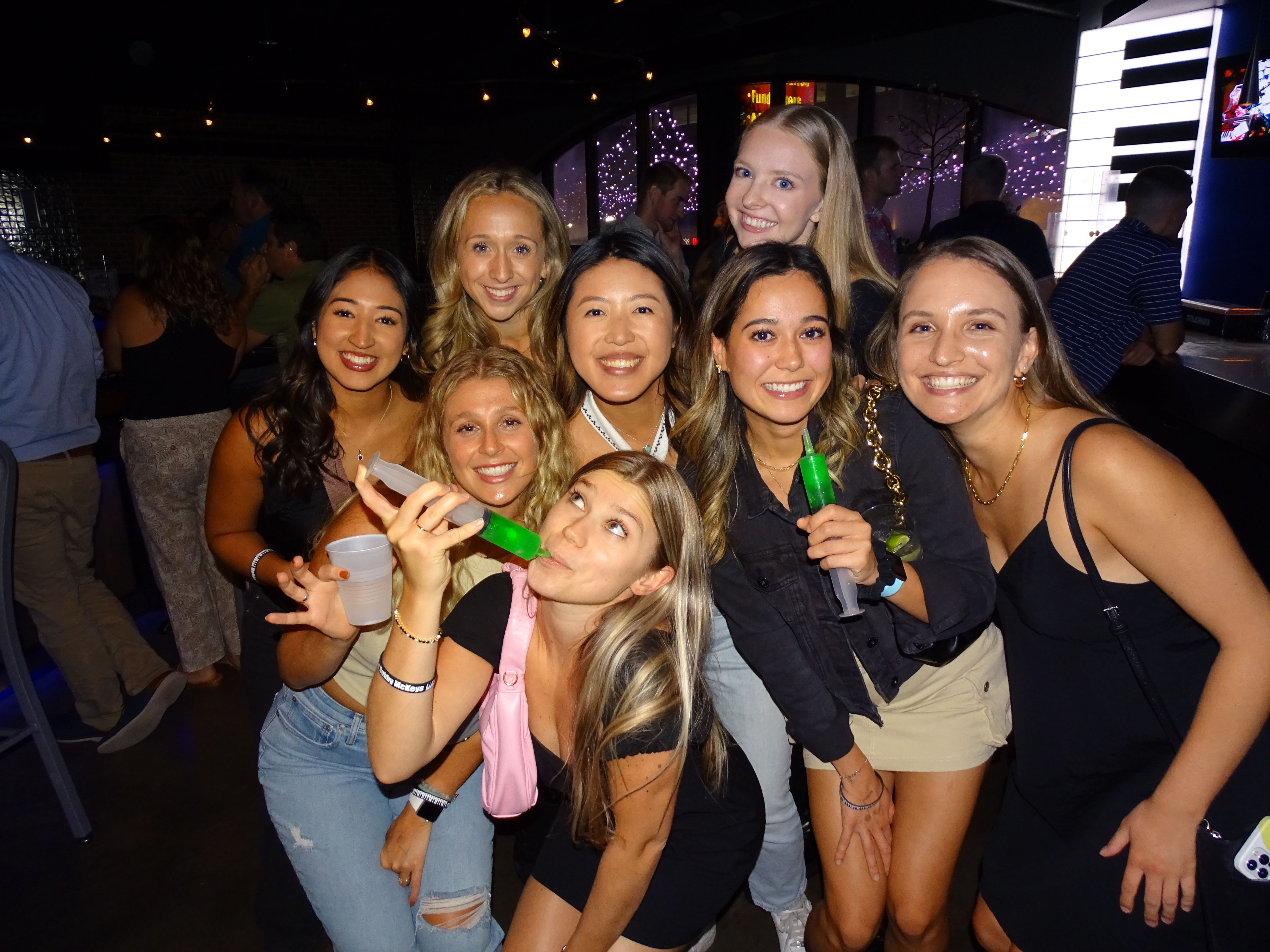 Group of nine friends smiling for a girls' night photo at a lively urban bar, holding neon green party shots and plastic cups under string lights and colorful wall decor.