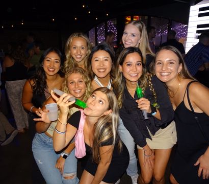 Group of nine friends smiling for a girls' night photo at a lively urban bar, holding neon green party shots and plastic cups under string lights and colorful wall decor.
