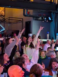Lively crowd at a packed indoor live-music venue cheering and singing, people with glow-necklaces and raised arms under neon and industrial lighting.