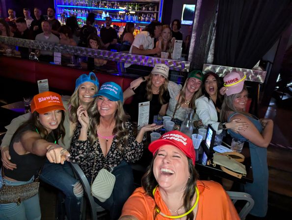 Group selfie of laughing women at a neon-lit bar wearing cheeky novelty hats and holding cocktails — lively girls' night/bachelorette party in a nightclub.