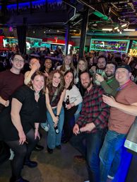 Large group of friends smiling and holding drinks in a crowded neon-lit downtown bar — lively nightlife scene with colorful backlit shelves and exposed ceiling