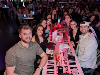 Smiling group of friends at a crowded nightlife venue, seated around a long piano-key patterned table with drinks and party decor — lively bar atmosphere
