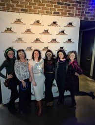 Bachelorette party group photo of six friends wearing festive glow glasses and a bride-to-be sash, posing in front of a brick-wall photo backdrop inside an indoor bar/event venue.
