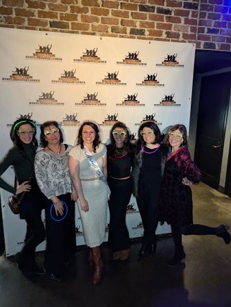 Bachelorette party group photo of six friends wearing festive glow glasses and a bride-to-be sash, posing in front of a brick-wall photo backdrop inside an indoor bar/event venue.
