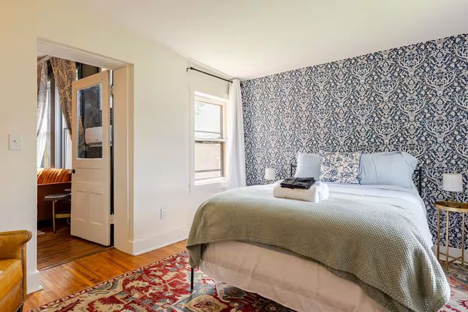 Cozy sunlit bedroom with a queen bed topped by a pale green throw and folded towels, blue damask accent wall, patterned area rug, wooden floors and bedside tables.