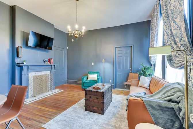 Cozy urban living room with blue-gray walls and hardwood floors, decorative fireplace with mounted TV, caramel leather sofa and teal armchair, patterned curtains, vintage trunk coffee table, chandelier and bright window light.