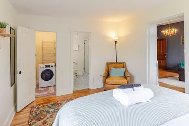 Bright cozy bedroom with made bed and folded towels, hardwood floors, mustard leather armchair, open doors showing a front-loading washer in a laundry closet and a tiled bathroom