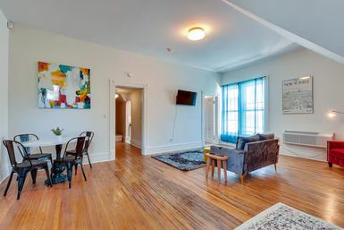 Bright open-concept apartment living room with polished hardwood floors, small dining nook with metal chairs and abstract wall art, cozy gray sofa and rug, wall-mounted TV and large window with blue curtains letting in natural light.