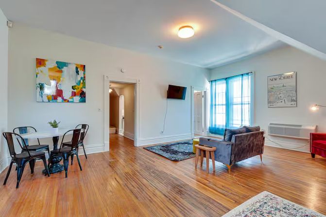 Bright open-concept apartment living room with polished hardwood floors, small dining nook with metal chairs and abstract wall art, cozy gray sofa and rug, wall-mounted TV and large window with blue curtains letting in natural light.