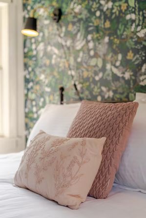 Close-up of two blush pink decorative pillows on white bed linens in a cozy bedroom with green floral wallpaper and a warm wall sconce