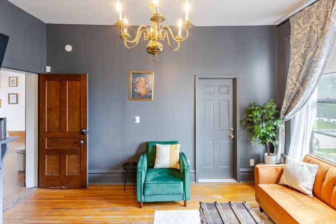 Cozy living room interior with gray walls, brass chandelier, emerald velvet armchair, burnt orange sofa, hardwood floors and a potted plant