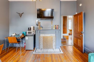 Cozy gray-walled apartment living room with hardwood floors, decorative fireplace and screen, wall-mounted TV, mini kitchenette with microwave and mini-fridge, orange dining chairs, and open wooden door