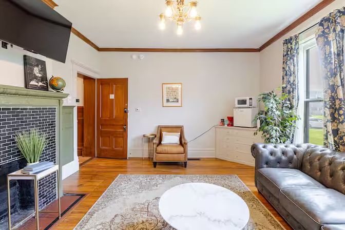 Cozy sunlit living room with tufted leather sofa, tan armchair, decorative fireplace, hardwood floors and patterned curtains.