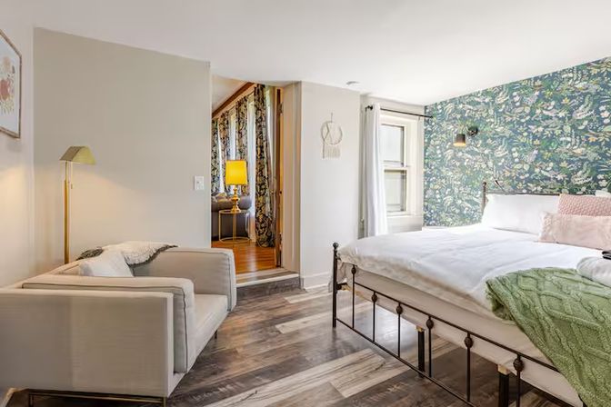 Bright, cozy apartment bedroom with a metal bed and white linens, green floral accent wall, hardwood floors, and a cushioned armchair reading nook by a floor lamp; doorway opens to a warmly lit living area.