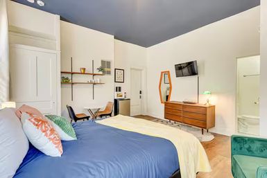 Bright modern studio bedroom with navy ceiling, queen bed dressed in blue duvet and colorful pillows, mid-century wooden dresser with wall-mounted TV, small dining table with chairs, open wall shelves and compact kitchenette, teal accent chair, hardwood floors and en-suite bathroom doorway