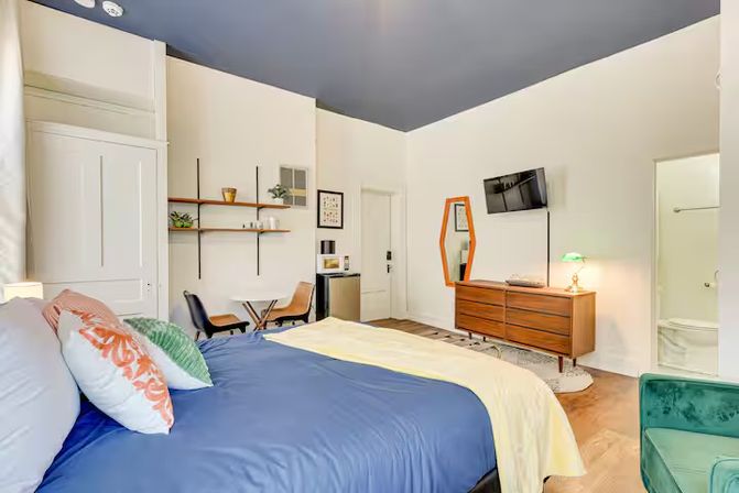 Bright modern studio bedroom with navy ceiling, queen bed dressed in blue duvet and colorful pillows, mid-century wooden dresser with wall-mounted TV, small dining table with chairs, open wall shelves and compact kitchenette, teal accent chair, hardwood floors and en-suite bathroom doorway