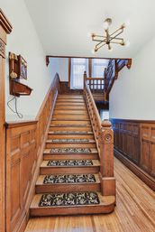 Historic home foyer with a grand wooden staircase and carved banister, floral-pattern stair runners on each step, wood-paneled wainscoting, a vintage wall telephone, and a modern brass chandelier.