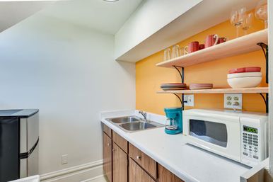 Compact kitchenette with cheerful orange accent wall, white countertop and double stainless sink, wooden cabinets, mini fridge, white microwave, teal coffee maker and open wooden shelves holding plates and glasses