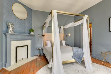Stylish bedroom with brass four‑poster canopy bed draped in white sheers, gray walls, decorative fireplace, hardwood floors and modern lamps.