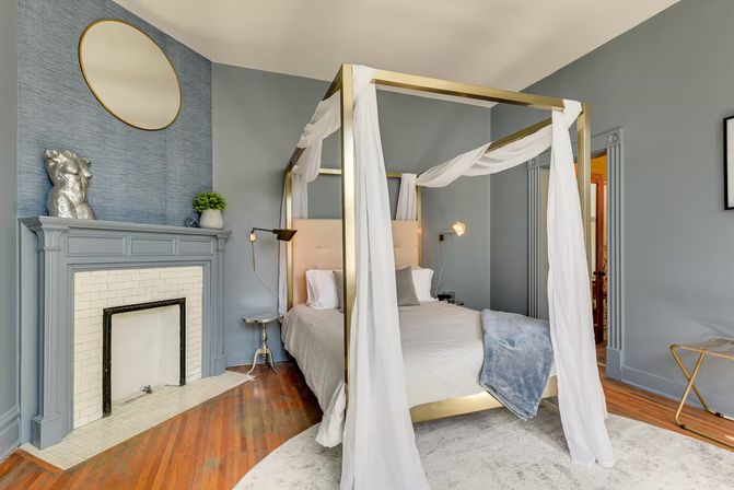 Stylish bedroom with brass four‑poster canopy bed draped in white sheers, gray walls, decorative fireplace, hardwood floors and modern lamps.