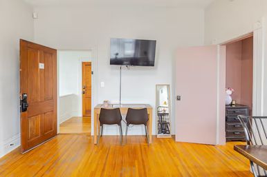 Bright apartment interior with polished hardwood floors, open wooden front door, wall-mounted TV above a small desk with two chairs, a full-length mirror leaning on the wall, and a partially open pink door to a bedroom.