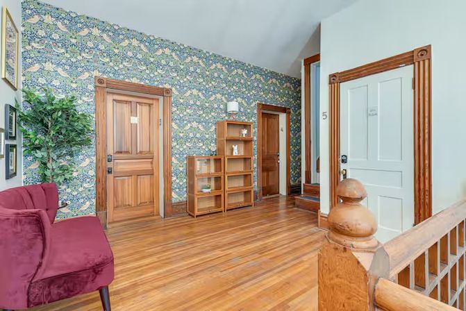 Bright historic-home hallway with polished hardwood floors, floral patterned wallpaper, carved wooden doors and trim, burgundy velvet bench, potted plant and open shelving.