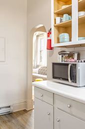 Bright apartment kitchen with white cabinets and countertop, stainless steel microwave, open shelves with pastel bowls, a red fire extinguisher by an arched doorway to a sunlit living room with tufted sofa and yellow ottoman