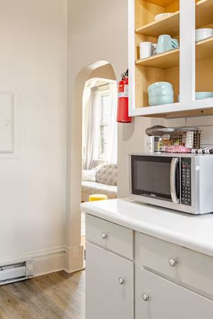 Bright apartment kitchen with white cabinets and countertop, stainless steel microwave, open shelves with pastel bowls, a red fire extinguisher by an arched doorway to a sunlit living room with tufted sofa and yellow ottoman