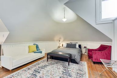Cozy loft-style attic bedroom with sloped ceiling, pendant light, queen bed with gray bedding, white sofa with yellow pillow, red velvet armchair, patterned area rug and hardwood floors.
