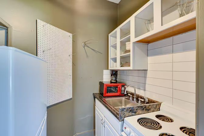 Cozy small kitchenette with white cabinets and subway-tile backsplash, stainless sink, coil electric stove, red microwave, coffee maker and pegboard on a gray wall.