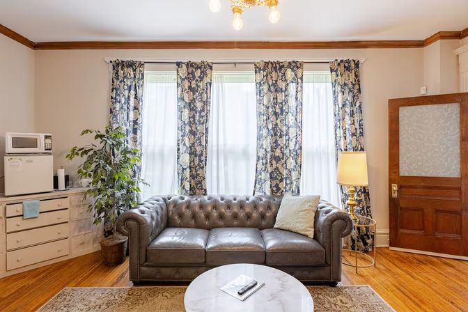 Cozy living room with tufted gray leather sofa, floral-pattern curtains on three tall windows, hardwood floors, potted plant, brass floor lamp and wooden door.