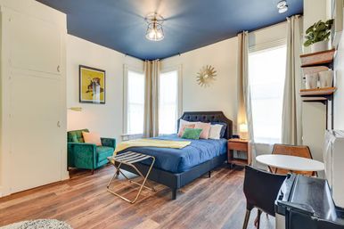 Cozy stylish studio bedroom with a navy-painted ceiling, queen bed in blue bedding, green velvet accent chair, wood floors, sunlit tall windows with curtains, small round dining table and sunburst wall decor