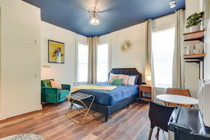 Cozy stylish studio bedroom with a navy-painted ceiling, queen bed in blue bedding, green velvet accent chair, wood floors, sunlit tall windows with curtains, small round dining table and sunburst wall decor