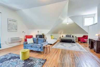 Bright attic loft apartment with sloped ceilings and hardwood floors, open living and sleeping area featuring a blue sofa, bed on a patterned rug, red accent chairs and a dormer window.
