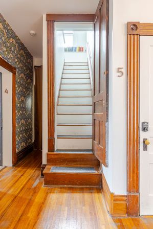 Cozy sunlit apartment hallway with narrow white stairs, worn wooden trim and hardwood floors, door number 5 and a small shelf of books at the top.