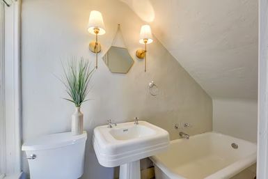Cozy small attic bathroom with sloped ceiling featuring a white pedestal sink, bathtub, toilet, hexagon mirror, brass wall sconces and a potted grass plant on the tank.
