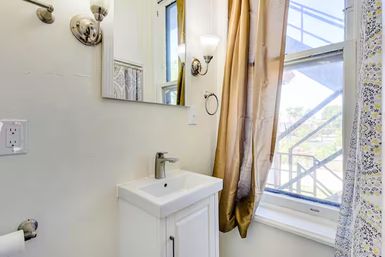 Bright small bathroom with white vanity sink and modern chrome faucet, wall mirror and sconce, gold window curtain and patterned shower curtain letting in natural light.