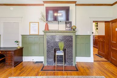 Bright period-style living room with green paneled wainscoting, black-tiled fireplace and mantel topped by a wall-mounted flat-screen TV, hardwood floors, wooden desk, and decorative plant and globe.