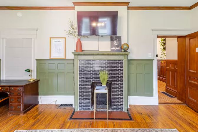 Bright period-style living room with green paneled wainscoting, black-tiled fireplace and mantel topped by a wall-mounted flat-screen TV, hardwood floors, wooden desk, and decorative plant and globe.