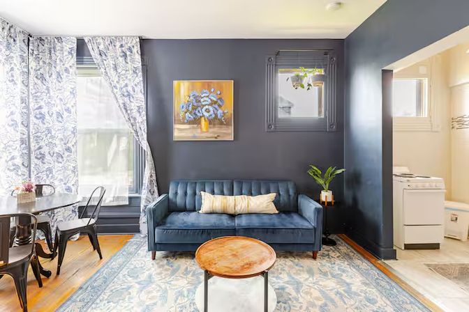 Cozy urban apartment living room with navy accent wall, blue velvet sofa, floral painting, patterned blue-and-white curtains, round wooden coffee table, dining nook and peek into compact kitchen.