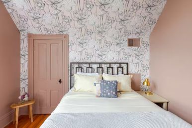 Cozy pink bedroom with sloped ceiling and black-and-white floral wallpaper, white bed with decorative pillows, brass bedside lamp and wooden stool