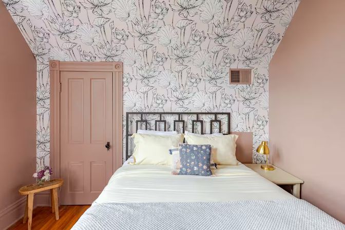 Cozy pink bedroom with sloped ceiling and black-and-white floral wallpaper, white bed with decorative pillows, brass bedside lamp and wooden stool