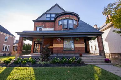 Historic red-brick Victorian house with a rounded turret, wide covered porch with hanging flower baskets, stone steps and a manicured front lawn under a clear blue sky.